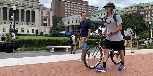 Person walking a bike while wearing a helmet on Columbia's Morningside campus near the Sundial on College walk.
