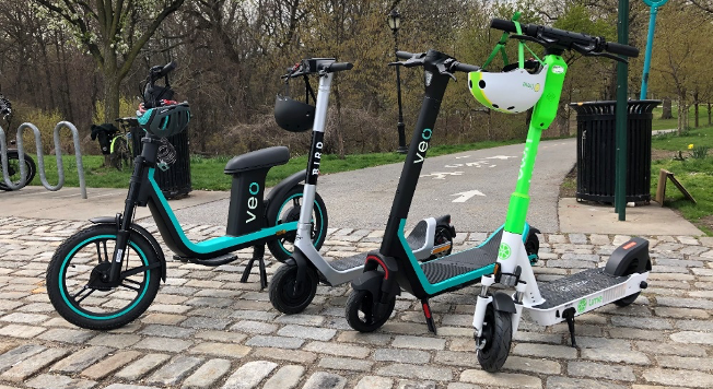 Four electric scooters on a sidewalk in a park entrance, each with a helmet dangling from the handlebars.