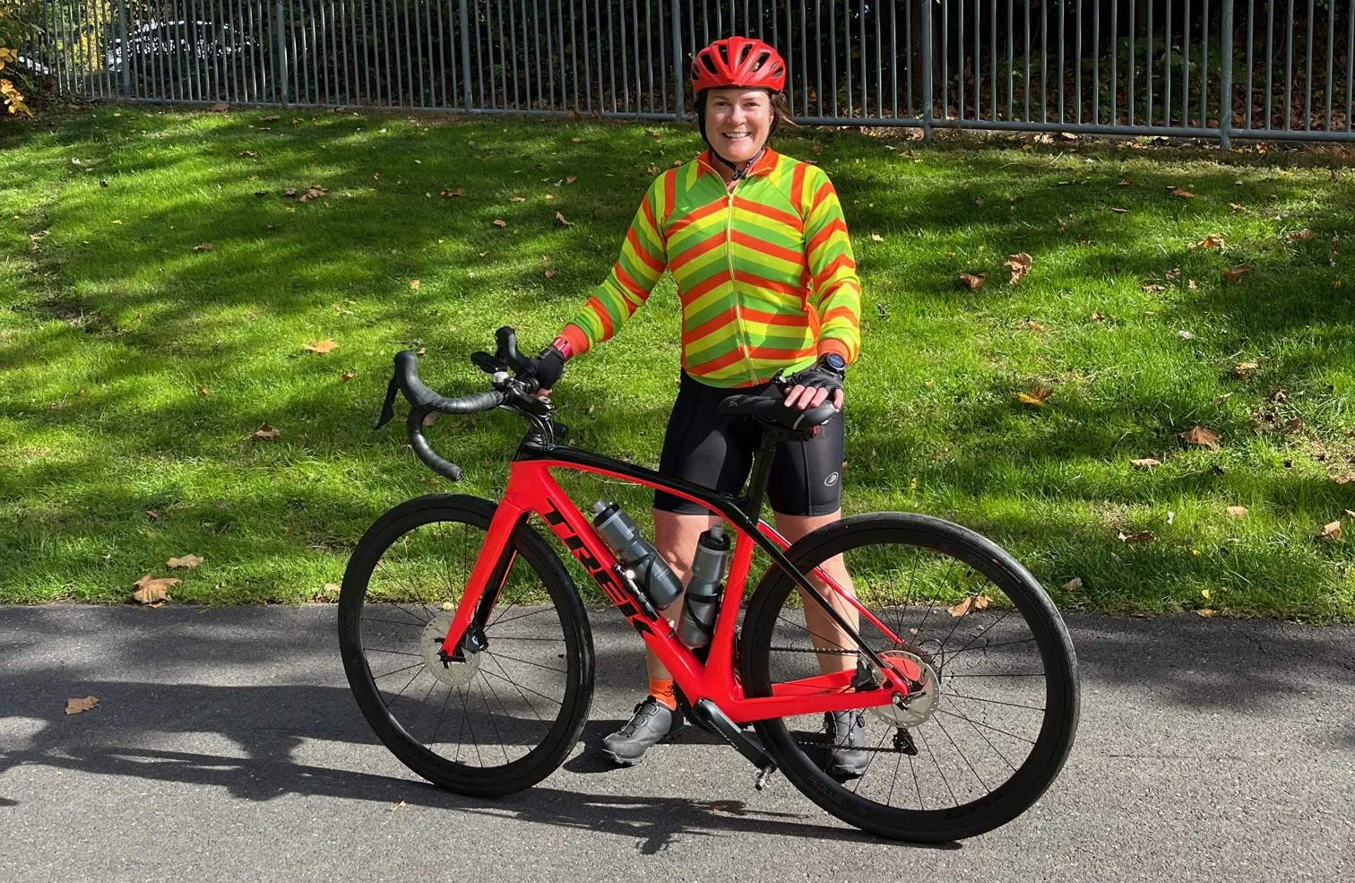 A woman wearing cycling clothes and a red helmet stands with her bicycle in front of green grass along her commute from New Jersey to Columbia